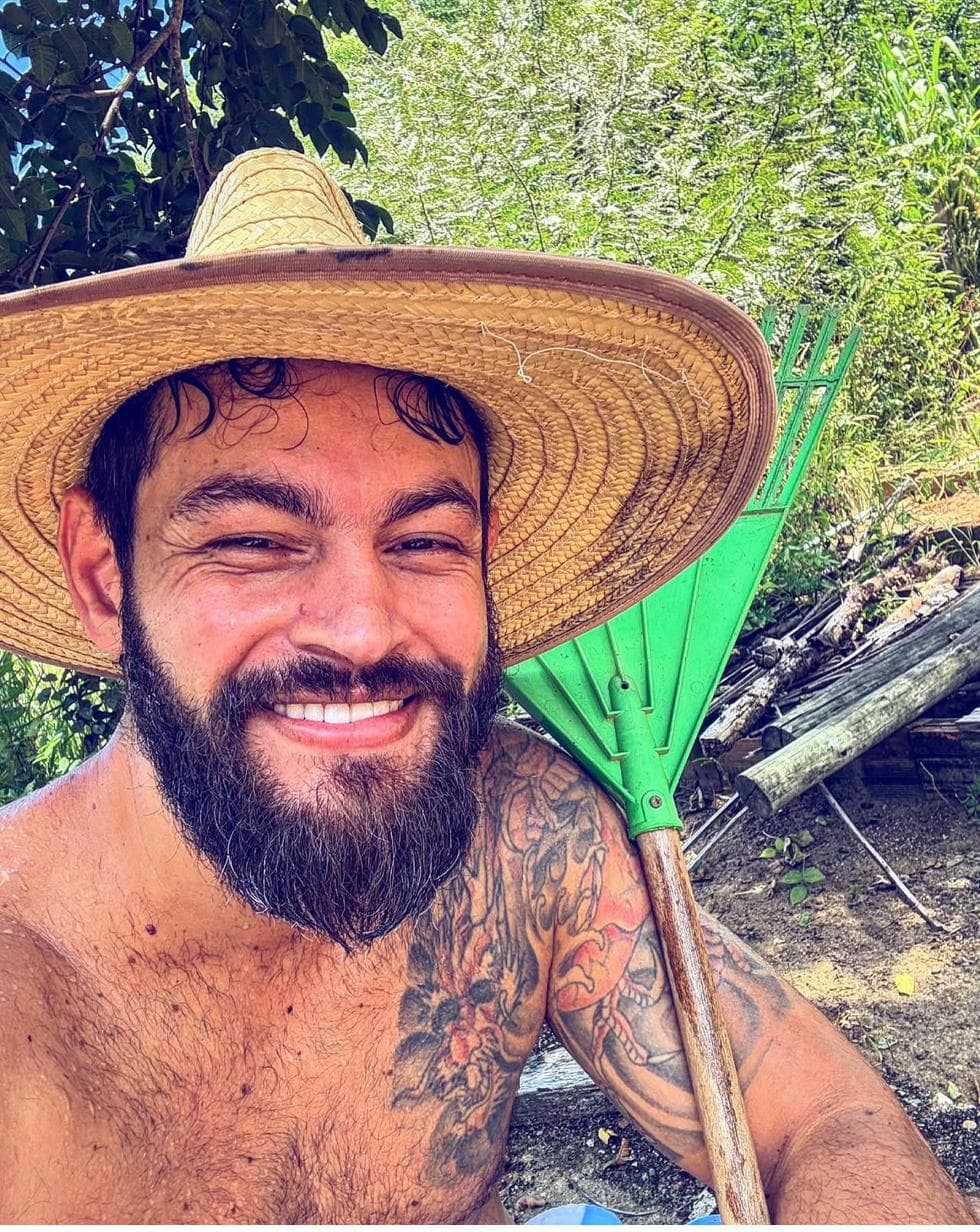 Thenperson sorrindo, de chapéu de palha, durante trabalho na roça.