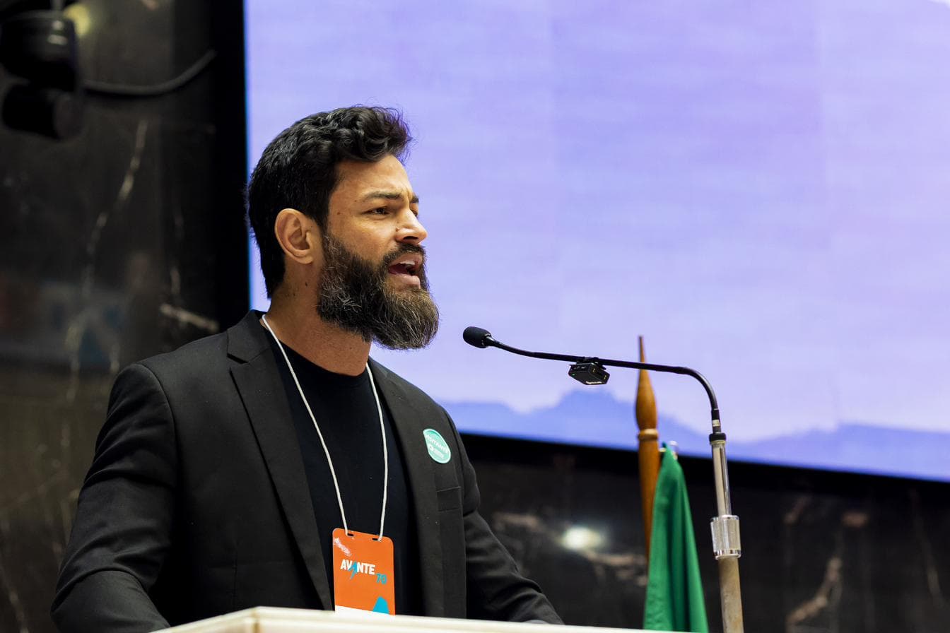 Thenperson discursando ao microfone em tribuna, visto de perfil, crachá do Avante 70 a tiracolo.